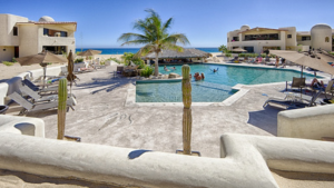 Pool area at a beachfront condo in Cabo, showing guests by the swim-up palapa bar and surrounding stucco buildings, framed by two tall cacti.