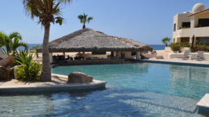 Resort swimming pool with a swim-up palapa bar, part of a beachfront vacation rental in Cabo San Lucas, with the beach and ocean in the background.