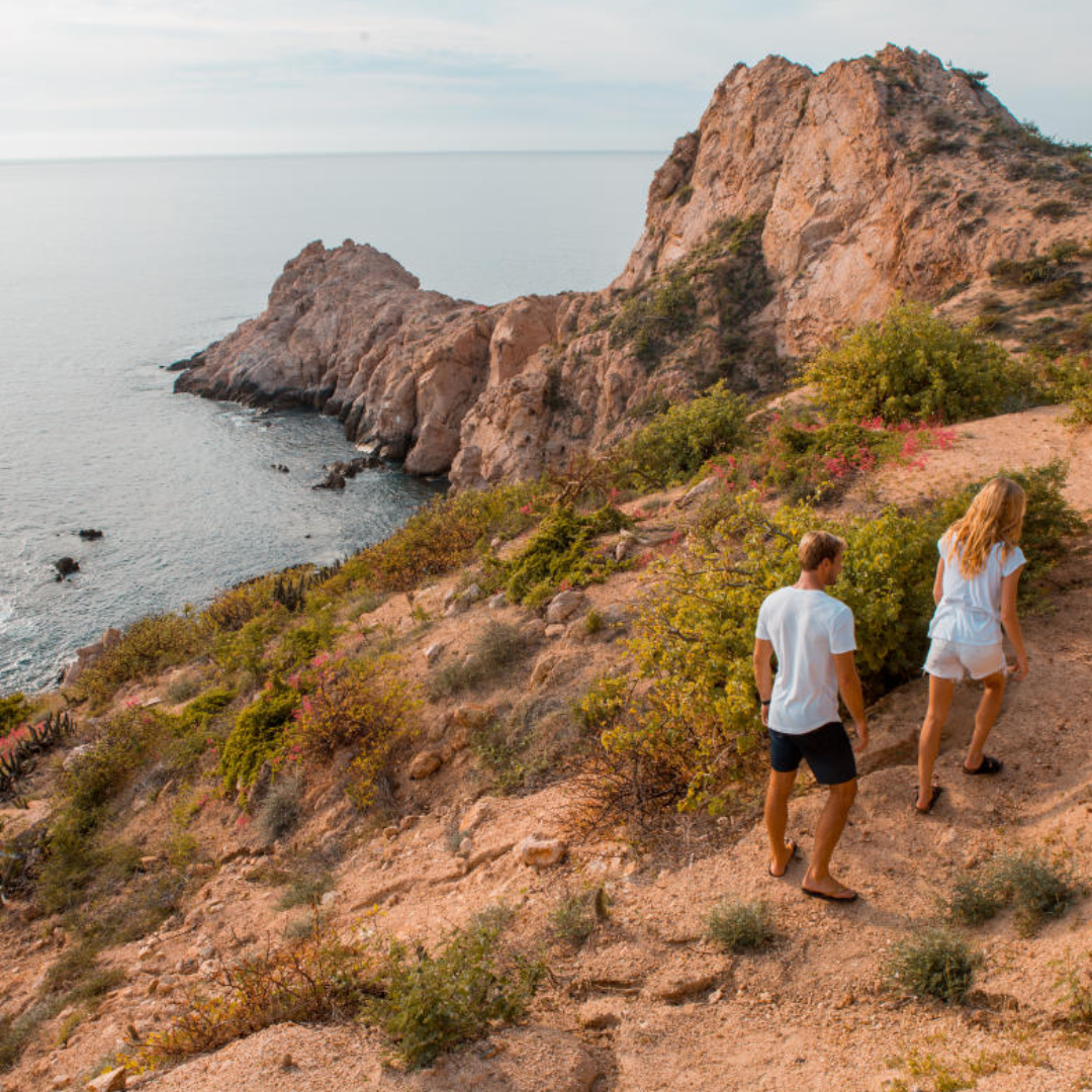 wellness-hiking-cabo A couple enjoying wellness hiking in Cabo on a scenic coastal trail overlooking the ocean and rocky cliffs.