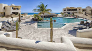 Pool area at a beachfront condo in Cabo, showing guests by the swim-up palapa bar and surrounding stucco buildings, framed by two tall cacti.