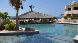 Resort swimming pool with a swim-up palapa bar, part of a beachfront vacation rental in Cabo San Lucas, with the beach and ocean in the background.
